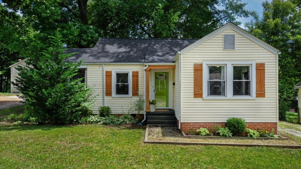 A small, single-story ranch or cottage-style house with light yellow or cream-colored horizontal siding and a dark grey shingled roof.